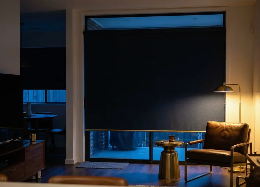Blockout roller blind fully lowered over a full-height sliding door, holding a living room dark at night